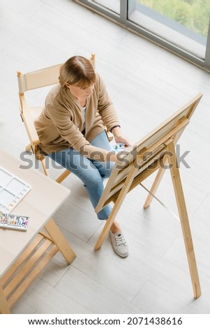 Top view shot of Caucasian old senior elderly retired female painter sitting on wooden chair using watercolor paintbrush and palette drawing painting art picture on canvas on easel stand in studio.