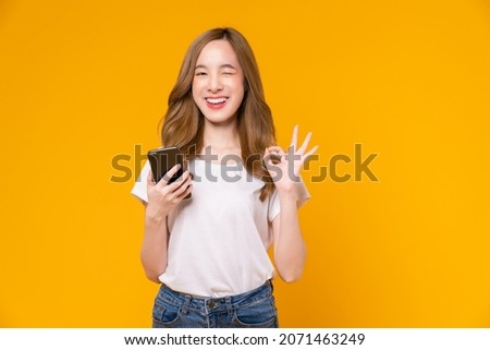 Cheerful Asian woman holding smartphone and shows ok sign on light yellow background. Royalty-Free Stock Photo #2071463249