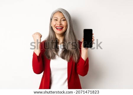 Success. Asian senior woman showing smartphone blank screen and fist pump, winning prize online, standing over white background