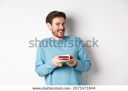 Excited man receive birthday surprise, holding bday cake and smiling happy, standing over white background, making wish on lit candle