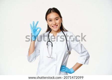 Smiling asian female physician, nurse in rubber gloves and medical uniform shows okay sign, approves and likes smth good, standing over white background