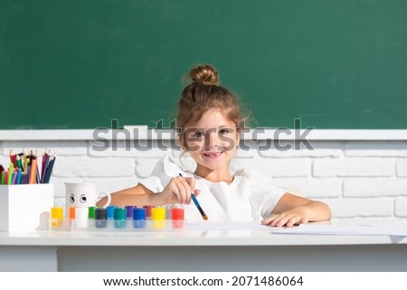 Painting school lesson, drawing art. Child girl draws in classroom sitting at a table, having fun on school blackboard background.