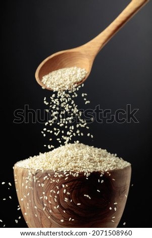 Grains of sesame are poured with a wooden spoon in the bowl. White sesame on a dark background. Copy space. Royalty-Free Stock Photo #2071508960