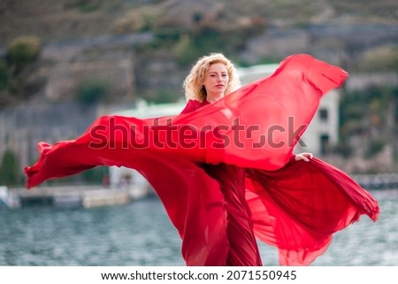 A woman in a red dress, a fashion model with long silk wings in a flowing dress, flying fabric on the embankment.