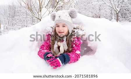 The girl lies in a snowdrift and smiles. The kid in a funny hat has built an igloo or a snow cave. Winter activity ideas for children. What to do for a child in winter. Ideas for winter outdoor games.