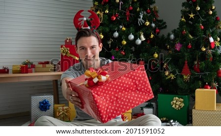 White Caucasian young man, good-looking in concept image, Christmas gift. The young man handed a gift box and looked at the camera. Stack of gift boxes and a Christmas tree decorated in the back.