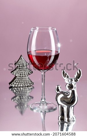 Glass of wine on a high thin leg on a pink background with a reflection, surrounded by a Christmas tree and a deer. Christmas menu. Vertical image. Space for copying.