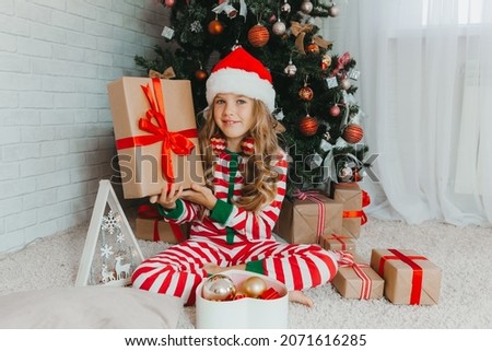 A little girl in a Santa hat sits on the floor near the Christmas tree and holds a gift in her hands. Christmas Eve.