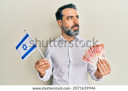 Middle age man with beard and grey hair holding israel flag and shekels banknotes smiling looking to the side and staring away thinking. 