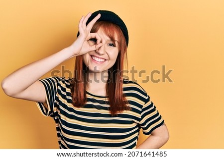Redhead young woman wearing wool cap smiling happy doing ok sign with hand on eye looking through fingers 