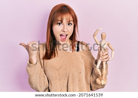 Redhead young woman holding small wooden manikin pointing thumb up to the side smiling happy with open mouth 