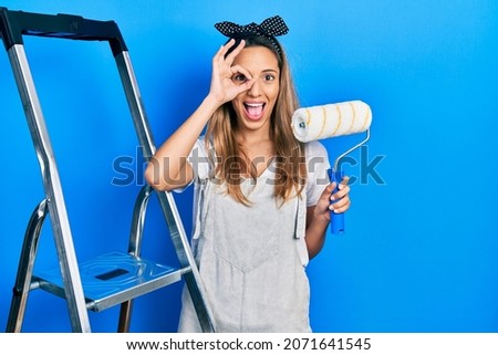 Beautiful hispanic woman by stairs holding roller painter smiling happy doing ok sign with hand on eye looking through fingers 