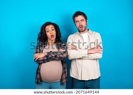 Shocked embarrassed young couple expecting a baby standing against blue background keeps mouth widely opened. Hears unbelievable novelty stares in stupor