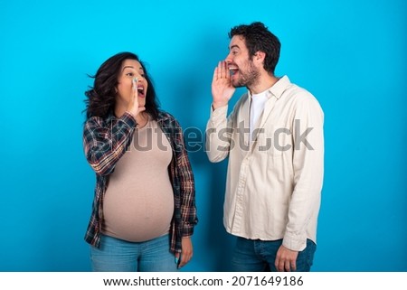 young couple expecting a baby standing against blue background look empty space holding hand near her face and screaming or calling someone.