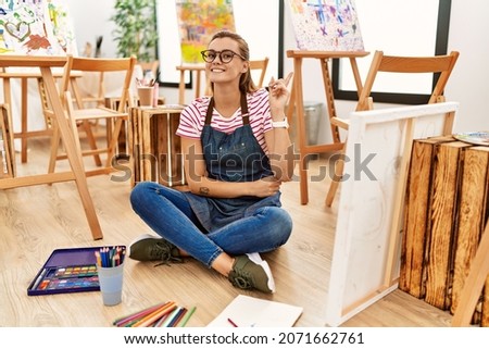 Young brunette woman at art studio sitting on the floor with a big smile on face, pointing with hand and finger to the side looking at the camera. 