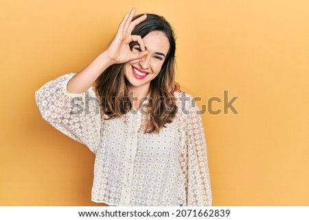 Young hispanic girl wearing casual clothes smiling happy doing ok sign with hand on eye looking through fingers 