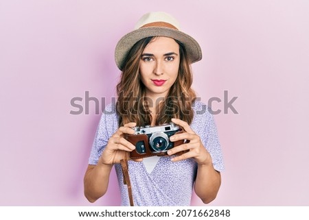Young hispanic girl wearing summer hat holding vintage camera relaxed with serious expression on face. simple and natural looking at the camera. 