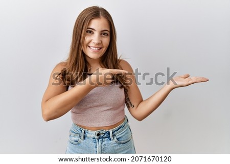 Young brunette woman standing over isolated background amazed and smiling to the camera while presenting with hand and pointing with finger. 