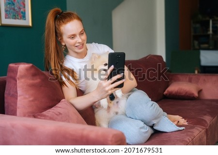 Happy redhead young woman cuddling pretty white small Spitz pet dog and taking selfie using mobile phone sitting on comfortable sofa, in cozy light living room with modern interior.