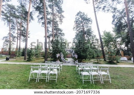 General plan of the registration area in the forest. Round flower arch among the trees. Wooden white chairs for guests. Wedding day and holiday atmosphere.