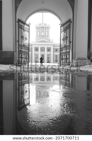 Black and white photograph of a manor house in Russia in winter.  Open gate.  The woman is walking alone.  Reflection in a snow puddle                               