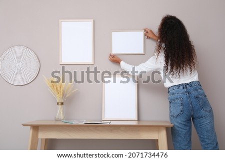 African American woman hanging empty frame on pale rose wall over table in room, back view. Mockup for design
