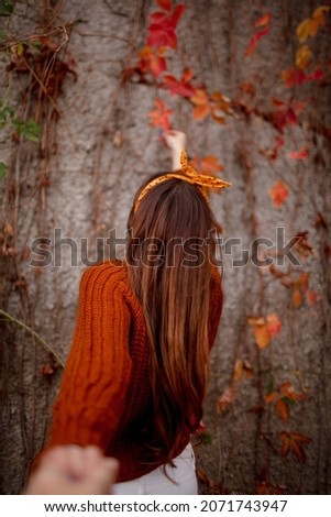 back view of woman dressed white trousers and brown sweater walking with pumpkin in the park against the fall leaves