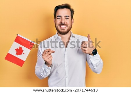 Handsome caucasian man with beard holding canada flag smiling happy and positive, thumb up doing excellent and approval sign 