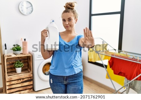Young caucasian woman holding detergent bottle at laundry room with open hand doing stop sign with serious and confident expression, defense gesture 