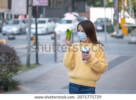 Beautiful Chinese Asian young woman walks on street during sunny autumn day. Holds drinks cup of coffee, takes pictures selfie using mobile cell phone. Wearing face mask to prevent respiratory disease