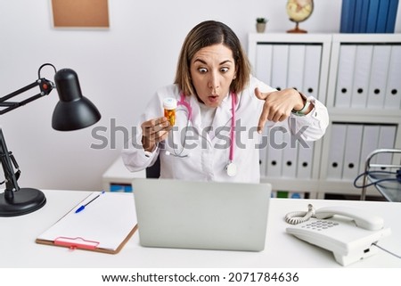 Young hispanic woman wearing doctor uniform holding pills at the clinic pointing down with fingers showing advertisement, surprised face and open mouth 