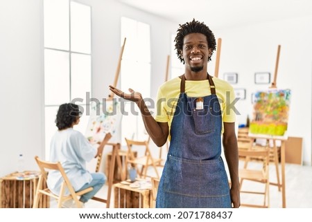 African young man standing at art studio smiling cheerful presenting and pointing with palm of hand looking at the camera. 