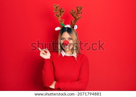 Beautiful hispanic woman wearing deer christmas hat and red nose with a big smile on face, pointing with hand and finger to the side looking at the camera. 