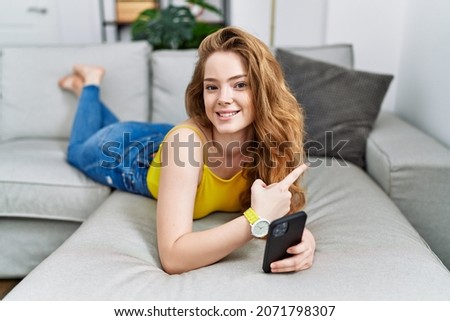 Young caucasian woman lying on the sofa using smartphone cheerful with a smile on face pointing with hand and finger up to the side with happy and natural expression 