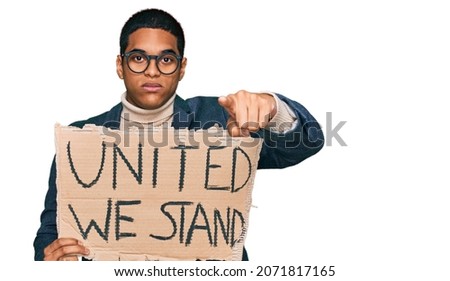 Young handsome hispanic man holding united we stand banner pointing with finger to the camera and to you, confident gesture looking serious 