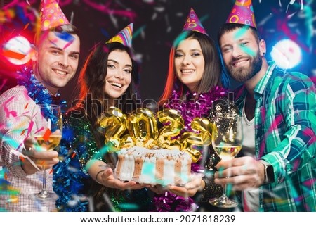 A group of young people in birthday caps with a cake in their hands with the numbers 2022 are looking at the camera against a dark background with light and music
