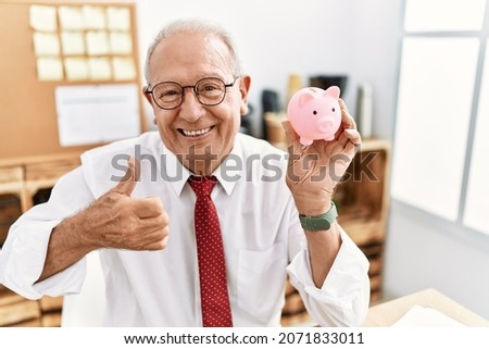 Senior business man holding piggy bank smiling happy and positive, thumb up doing excellent and approval sign 