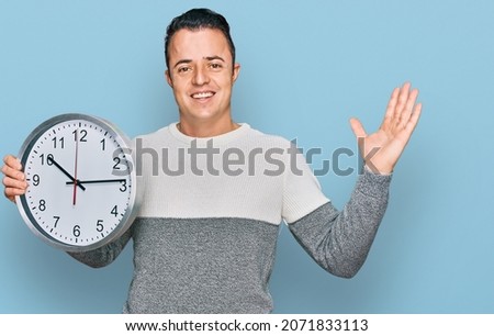 Handsome young man holding big clock celebrating victory with happy smile and winner expression with raised hands 