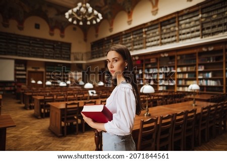 Portrait of attractive woman in white clothes with books in hands in beautiful public library posing at camera with serious face. Female student reads books in the student library.