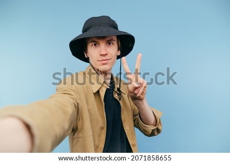 Cute young man in panama taking selfie on smartphone and showing peace gesture on blue background.