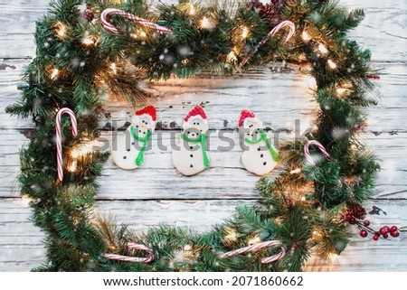 Holiday wreath made of Christmas garland with three Iced Christmas Snowman cookies over a white rustic wood background. Image shot from flat lay or top view position.