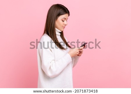 Side view portrait of female using smartphone, checking social networks, has concentrated expression, wearing white casual style sweater. Indoor studio shot isolated on pink background.