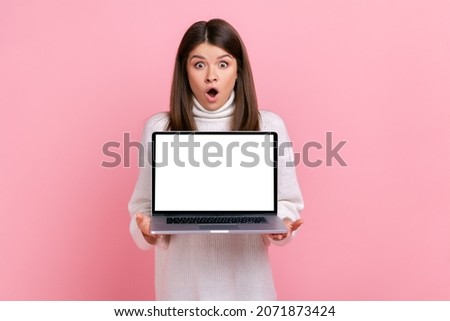 Astonished female holding notebook with empty display in hands, looking at camera with open mouth, wearing white casual style sweater. Indoor studio shot isolated on pink background.