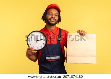 Food delivery, shipping on time! Courier man wearing overalls and T-shirt holding clock and parcel, express courier transportation, post mail services. Indoor studio shot isolated on yellow background