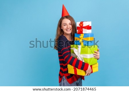 Smiling woman wearing striped casual style sweater, celebrating birthday, embracing present boxes, looking at camera with satisfied expression. Indoor studio shot isolated on blue background.