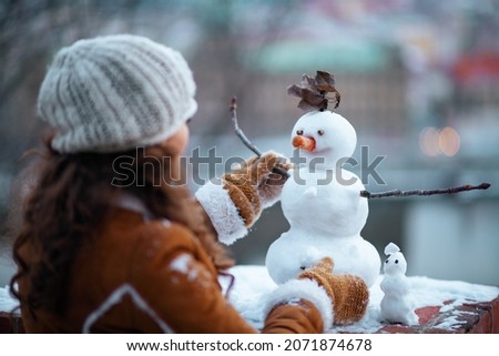 Seen from behind middle aged woman with mittens in a knitted hat and sheepskin coat making a snowman outside in the city park in winter. Royalty-Free Stock Photo #2071874678