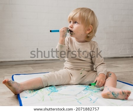 A little girl in pajamas is sitting on the floor and dreamily gnawing on a water marker. The face and clothes are stained with paint