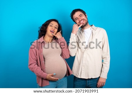 young couple expecting a baby standing against blue background smiling doing phone gesture with hand and fingers like talking on the telephone. Communicating concepts.