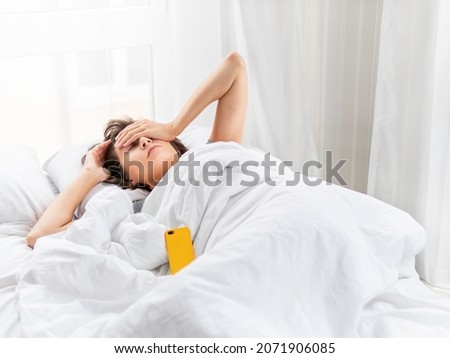 Sleepy woman is lying in bed, completely covered with white blanket. Smartphone used as alarm clock. It's hard to wake up early in morning. Woman does not get enough sleep. Royalty-Free Stock Photo #2071906085