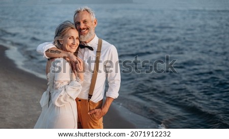 Senior newlyweds hug. Stylish couple of elderly newlyweds stand embracing on river bank. Royalty-Free Stock Photo #2071912232